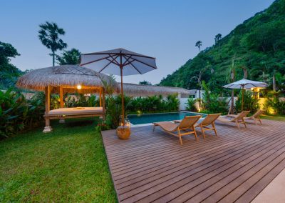 Dusk backyard at Aman Amed at Vesica Villas Resort in Amed, with four loungers on a wood deck, white umbrella, lit thatched bale on the lawn beside the pool, tropical planting, and a steep green hillside under twilight sky