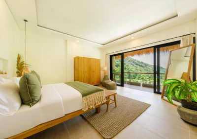 Bedroom at Aman Amed at Vesica Villas Resort in Amed, angled toward open balcony doors over a green hillside, with olive bed accents, wardrobe, grey beanbag, leaning wood mirror, and a potted plant