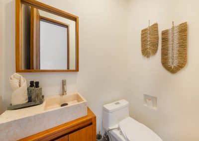 Powder room at Aman Amed at Vesica Villas Resort in Amed, with a stone vessel sink on a wood vanity, square wood-framed mirror, white toilet with recessed paper niche, and leaf-shaped woven wall hangings
