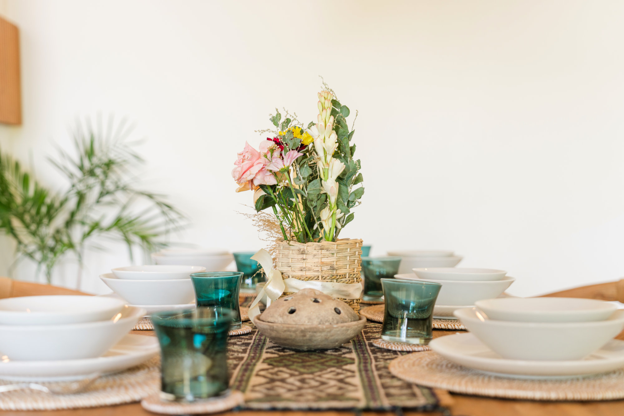 Aman Amed Balcony Dining table laid with neutral colored plates and dished with teal tinted glasses and a flower arrangement in the enter of the table.