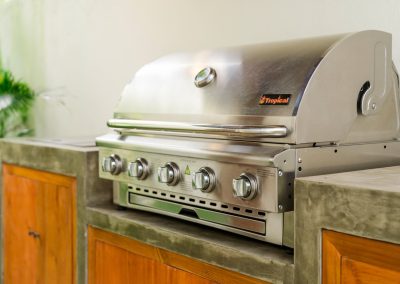 Built-in stainless steel gas grill on a grey concrete outdoor kitchen counter at Aman Amed at Vesica Villas Resort in Amed, with wood cabinets below and tropical foliage beside the patio wall