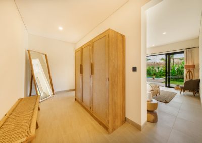 Dressing area at Aman Amed at Vesica Villas Resort in Amed, with a woven bench, leaning wood floor mirror, three-door wood wardrobe, and open doorway into a bedroom with garden view through glass doors
