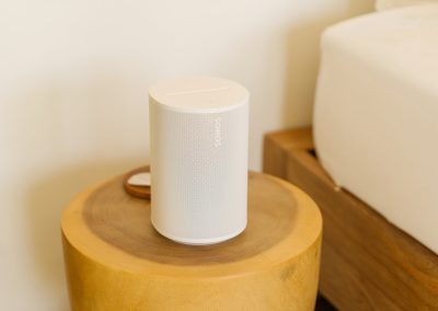 White smart speaker on a cylindrical solid-wood nightstand beside a white-linen bed at Aman Amed villa at Vesica Villas Resort in Amed, against a plain off-white wall in soft natural light