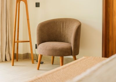 Rounded brown upholstered armchair on light tile at Aman Amed villa at Vesica Villas Resort in Amed, beside a slim wood floor-lamp stand, cream curtain, and soft natural light