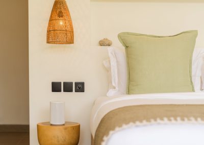 Bedside corner at Aman Amed at Vesica Villas Resort in Amed, with a sage green pillow, olive throw, conical woven pendant, black wall switches, and a round wood stump table holding a white speaker