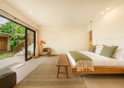 Bedroom at Aman Amed at Vesica Villas Resort in Amed, with a white bed and olive accents, woven bench, grey armchair and tripod lamp, open black-framed doors to a garden path, carved door, and outdoor stair