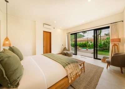 Bedroom looking out to the pool deck at Aman Amed at Vesica Villas Resort in Amed, with white bed and olive accents, woven pendant, grey armchair, wood-slat door, and open doors to loungers, umbrella, and thatched bale