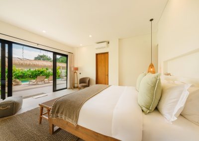 Bedroom opening to the pool patio at Aman Amed at Vesica Villas Resort in Amed, with white bed and sage pillows, brown woven throw, grey armchair, and open glass doors to loungers by the private pool and thatched roof