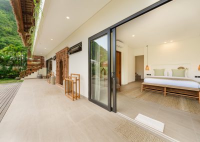 Bedroom terrace at Aman Amed at Vesica Villas Resort in Amed, with open sliding doors to a white bed with sage pillows, outdoor towel rack, carved wood wall panel, stair under thatch, and forested hillside beyond