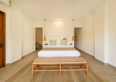 Centered bedroom at Aman Amed at Vesica Villas Resort in Amed, with white linens, sage pillows and tan runner, woven bench on a jute rug, twin conical woven pendants, flanking doorways, and a wood door to the left