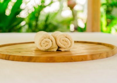 Two rolled cream towels on a round wooden tray at Aman Amed villa at Vesica Villas Resort in Amed, set on a white surface with soft bokeh of tropical greenery and sunlight beyond