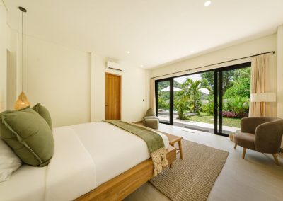 Bedroom at Umah Sukah at Vesica Villas Resort in Amed with a wooden bed in white linens and sage pillows, grey armchair, jute rug, and large glass doors framing a garden, white umbrella, and distant hillside