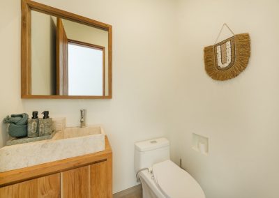 Powder room at Umah Sukah at Vesica Villas Resort in Amed with a floating wood vanity, cream stone vessel sink, tall wood-framed mirror, modern white toilet, and a circular fringed natural-fiber wall hanging