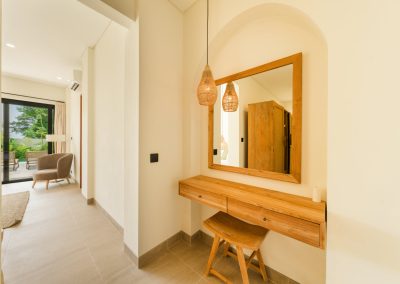 Dressing vanity at Umah Sukah at Vesica Villas Resort in Amed with a floating wood desk and stool in an arched niche, square wood-framed mirror, teardrop rattan pendant, and a glimpse of the bedroom terrace beyond