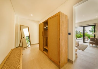 Dressing hallway at Umah Sukah at Vesica Villas Resort in Amed with an open three-section wood wardrobe, leaning full-length mirror, rattan-seat bench, and a living space opening to the turquoise pool and tropical greenery