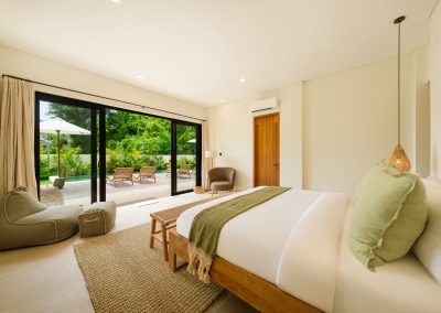 Bedroom suite at Umah Sukah at Vesica Villas Resort in Amed opening to the private pool deck, with white bedding and sage accents, olive beanbag on a jute rug, grey armchair, and wooden loungers under a white umbrella outside