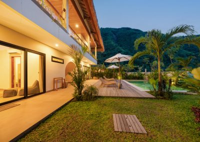 Twilight exterior of Umah Sukah at Vesica Villas Resort in Amed with warm balcony lighting, wooden pool deck, loungers under a white umbrella, manicured lawn, and a steep green mountain rising behind the two-story villa