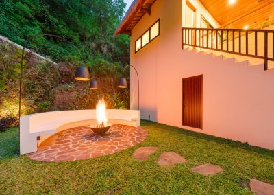 Night fire-pit circle at Umah Sukah at Vesica Villas Resort in Amed with roaring flames in a metal bowl on red stone paving, curved white bench, black arc lamps, ivy-covered hillside, and warm villa lights under the thatch
