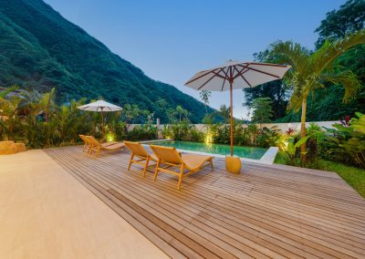 Dusk pool deck at Umah Sukah at Vesica Villas Resort in Amed with wooden loungers under white umbrellas, turquoise rectangular pool, tropical planting with warm uplights, and a steep green mountain against twilight sky