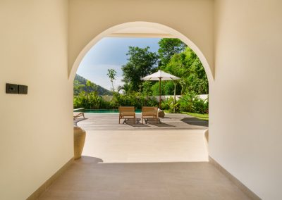 View through a white plaster archway at Umah Sukah at Vesica Villas Resort in Amed to the sunlit terrace, wooden loungers, white umbrella, turquoise pool, and lush hillside framed by cream interior walls