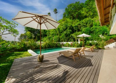 High-angle pool deck at Umah Sukah at Vesica Villas Resort in Amed with turquoise water, weathered wood planks, timber loungers under white umbrellas, thatched roof corner, and a steep tropical hillside under blue sky