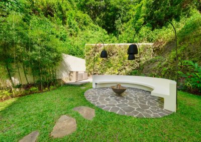 Garden fire-pit seating at Umah Sukah at Vesica Villas Resort in Amed with white curved bench, dark irregular stone circle, metal fire bowl, three black arched lamps, bamboo thicket, and a steep forested hillside