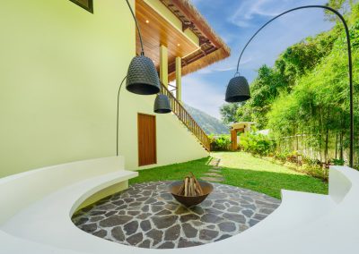 Outdoor fire-pit court at Umah Sukah at Vesica Villas Resort in Amed with white semicircular bench, crazy-pave stone patio, log-filled metal bowl, three black pendant arc lamps, wooden stair, and green hills under blue sky