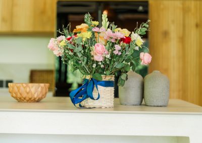 Kitchen island floral vignette at Umah Sukah at Vesica Villas Resort in Amed with pink roses and yellow blossoms in a woven basket tied with a blue ribbon, grey ceramic bud vases, and soft-focus wood cabinetry behind