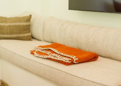 Daybed seating detail at Umah Sukah at Vesica Villas Resort in Amed with cream linen cushions, a terracotta throw edged in white whipstitch, olive striped accent pillow, and the edge of a wall-mounted TV above