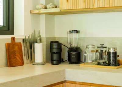 Kitchen counter corner at Umah Sukah at Vesica Villas Resort in Amed with Nespresso machine, Philips blender, milk frother, French press, magnetic knife strip, cutting board, and vertical-slat wood cabinets under a floating shelf