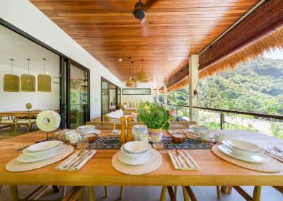 Set wooden dining table on the terrace at Umah Sukah at Vesica Villas Resort in Amed with white ceramics on woven mats, floral centerpiece, polished wood ceiling and fan, glass railing over a forested hillside, and lounge seating beyond