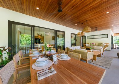 Covered terrace dining and living at Umah Sukah at Vesica Villas Resort in Amed with a fully set wood table, flower vase, rattan chairs, cream sofa lounge under tiered wicker pendants, wood-plank ceiling fans, and garden greenery