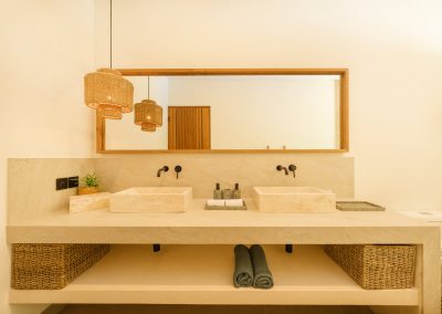 Double vanity bathroom at Umah Sukah at Vesica Villas Resort in Amed with twin stone vessel sinks, black wall faucets, wide wood-framed mirror, woven rattan pendants, open shelf with baskets and teal towels, and a glimpse of the toilet