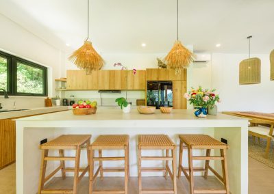 Kitchen island at Umah Sukah at Vesica Villas Resort in Amed with four wood bar stools and rattan seats, fruit bowl and floral arrangement, shaggy straw pendants overhead, black sink faucet by a garden window, and dining beyond