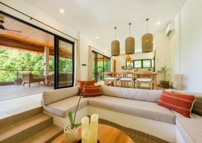 Open living and dining at Umah Sukah at Vesica Villas Resort in Amed with a sunken beige sofa and terracotta pillows around a wood coffee table, dining under cylindrical wicker pendants, and open doors to a wood-ceiling jungle balcony