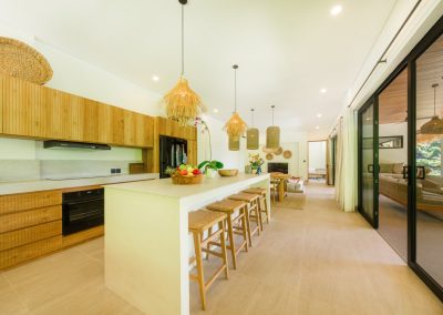 Kitchen looking into living at Umah Sukah at Vesica Villas Resort in Amed, with white island and wood stools, vertical-slat cabinetry, black oven and range hood, tiered woven pendants, and glass doors reflecting the bright tropical interior