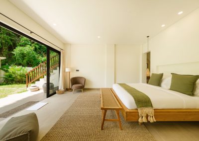 Bright bedroom at Umah Sukah at Vesica Villas Resort in Amed with a wooden platform bed in white linens and sage pillows, a wood bench on a jute rug, rounded armchair with floor lamp, and open black-framed doors to the tropical garden