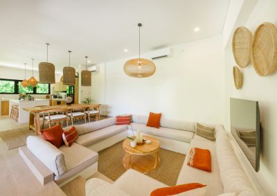 Living looking through dining to kitchen at Umah Sukah at Vesica Villas Resort in Amed, with sunken cream sofa and orange pillows, round wood table under an oval rattan pendant, wood dining with cylinder lights, and kitchen windows to tropical greenery