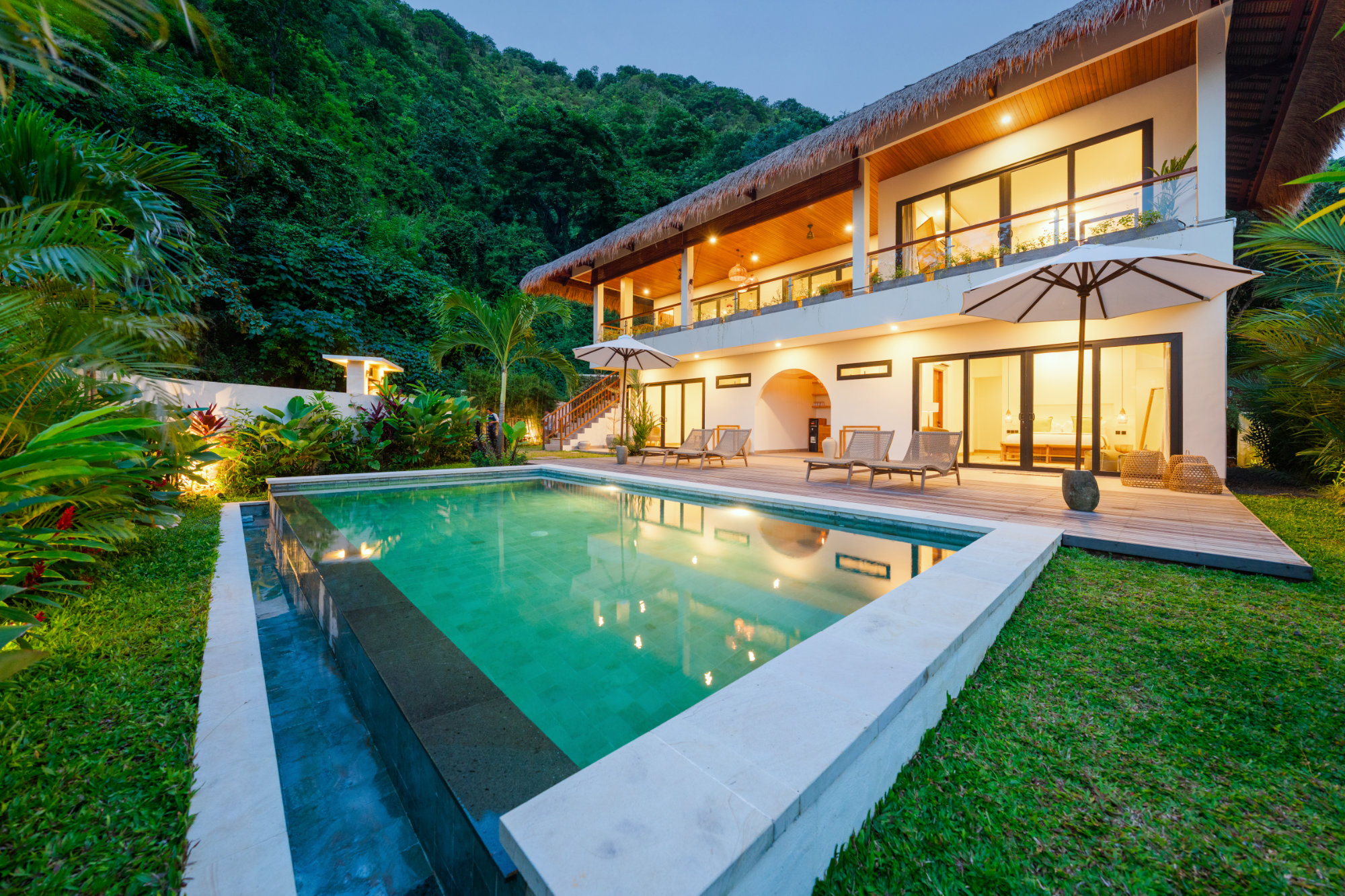 Twilight poolside at Umah Sukah at Vesica Villas Resort in Amed with glowing turquoise pool, dark wood deck and loungers under white umbrellas, warmly lit two-story thatched villa, manicured lawn, and dense green hillside