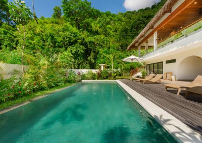Daytime private pool at Umah Sukah at Vesica Villas Resort in Amed with turquoise water, wood loungers on a dark deck, white umbrella, arched villa entry and thatched upper balcony, and a lush tropical hillside under blue sky