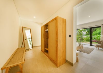 Dressing hallway at Umah Sukah at Vesica Villas Resort in Amed with a light-wood rattan-seat bench, leaning full-length wood mirror, open freestanding wardrobe, and a view into a sunlit bedroom opening to the garden