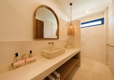 Ensuite vanity at Villa Svara with a fluted cream stone sink, arched wood mirror, woven raffia pendant, pink rolled towels, brass faucet, and herringbone-tiled shower beyond