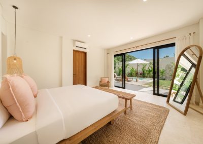 King bedroom at Villa Svara opening to the pool terrace, with white bedding and pink pillows, wood bench, oval floor mirror, wooden armchair, marble floor, and tropical garden beyond