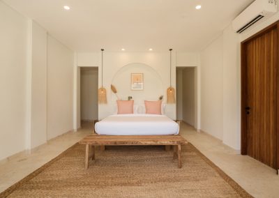 Centred king bedroom at Villa Svara with white linens and pink pillows, recessed arched niche, twin woven rattan pendants, rustic wood bench on a jute rug, and marble-tiled floor