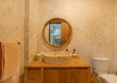 Bathroom vanity at Villa Svara at Vesica Villas Resort in Amed, with a cream stone vessel sink, circular wood-framed mirror, beige mosaic tile walls, pink waffle towel, and marble floor