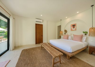 King bedroom at Villa Svara at Vesica Villas Resort in Amed, with white linens and pink pillows, rattan pendant lights, a wood bench on a jute rug, and black-framed doors to a tropical patio