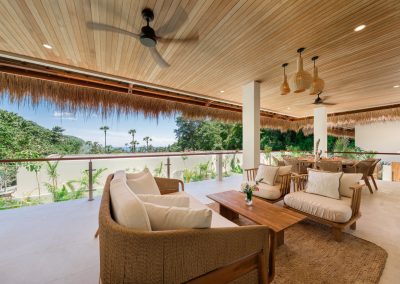 Upper terrace at Villa Svara at Vesica Villas Resort in Amed, with a woven lounge set on a jute rug, round wood dining table, rattan pendants, wood-slat ceiling, and a glass rail overlooking palms and sea