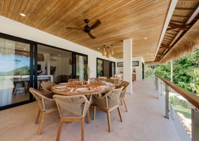 Outdoor dining terrace at Villa Svara at Vesica Villas Resort in Amed, with a round wood table set with pink napkins and amber glasses, woven chairs, marble floors, and a wood-slat ceiling