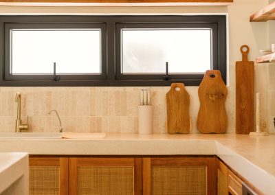 Kitchen counter at Villa Svara at Vesica Villas Resort in Amed, with brushed brass faucets, organic wood cutting boards, rattan-inlay cabinets, and black-framed windows over a cream stone top