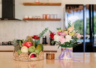 Kitchen welcome display at Villa Svara at Vesica Villas Resort in Amed, with a woven fruit basket, rose bouquet, and candle on a cream counter before floating shelves and a black fridge
